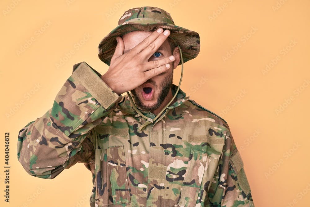 Young caucasian man wearing camouflage army uniform peeking in shock covering face and eyes with hand, looking through fingers afraid