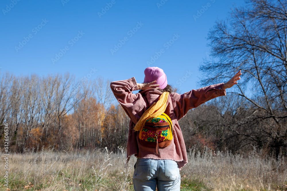 Mujer morena joven sosteniendo gorro mientras está de pie con la mano levantada en el campo durante una excursión