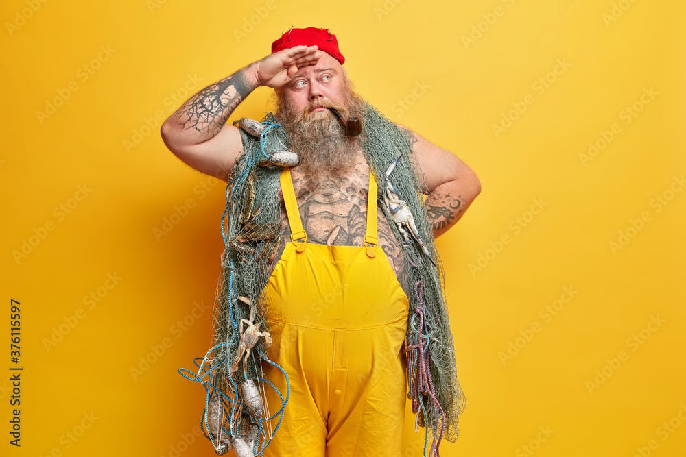 Thoughtful boatswain keeps hand on forehead, looks into distance while travels by ship, carries fishing net, dressed in yellow overalls, smokes pipe, stands indoor. Pensive fisherman at deck.
