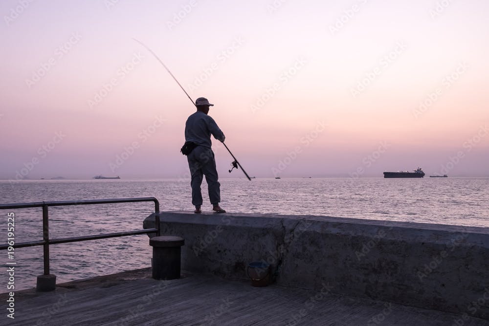 Elderly man silhouette standing on stone pier and throwing a fishing rod into the sea on beautiful sunset background. Boats and remote islands in haze on horizon line.