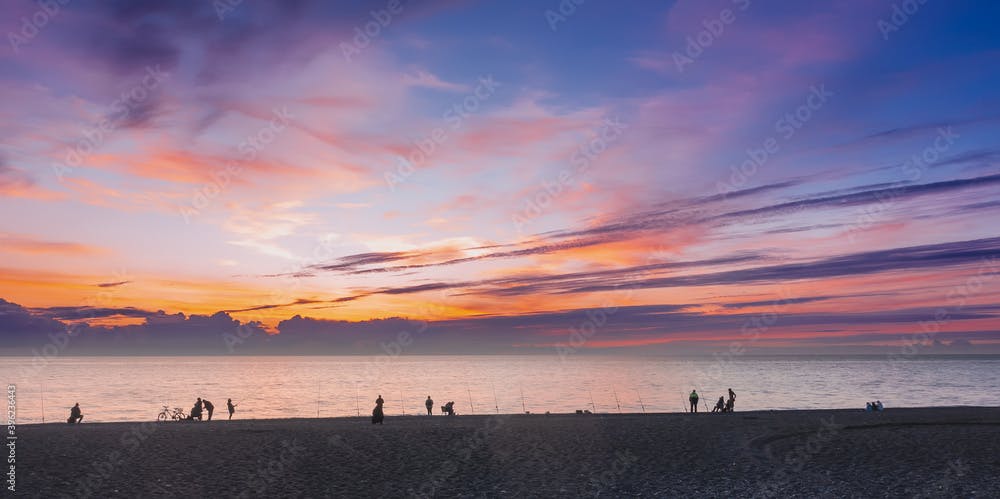 After sunset, the sky is colored in orange, scarlet, pink, lilac shades. Silhouettes of people and fishing rods are visible on the seashore. Evening fishing. Russia. Sochi