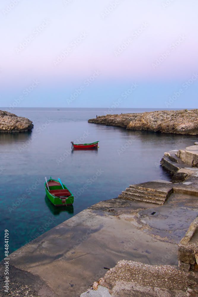 Polignano a Mare (BA): coast of rocks at violet/blu sunset. Some gozzo (fish boats) at anchor