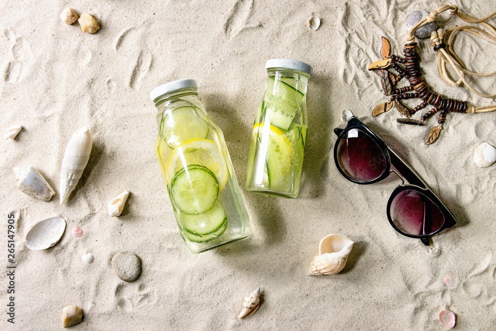 Summer theme. Two glass bottles with lemon and cucumber infusion sassy water, shells, sea stones, sunglasses, wooden beads, on white sand as background. Flat lay