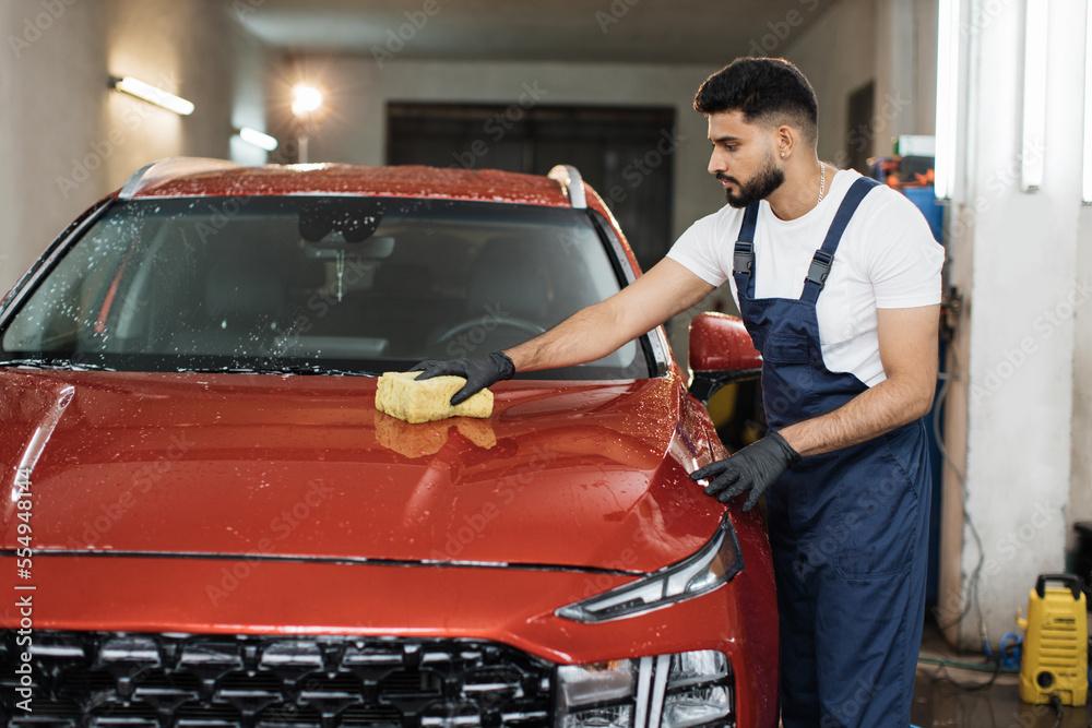 Car washing and detailing concept. Young concentrated bearded male worker holding the sponge in hand and cleaning red car hood with soap.