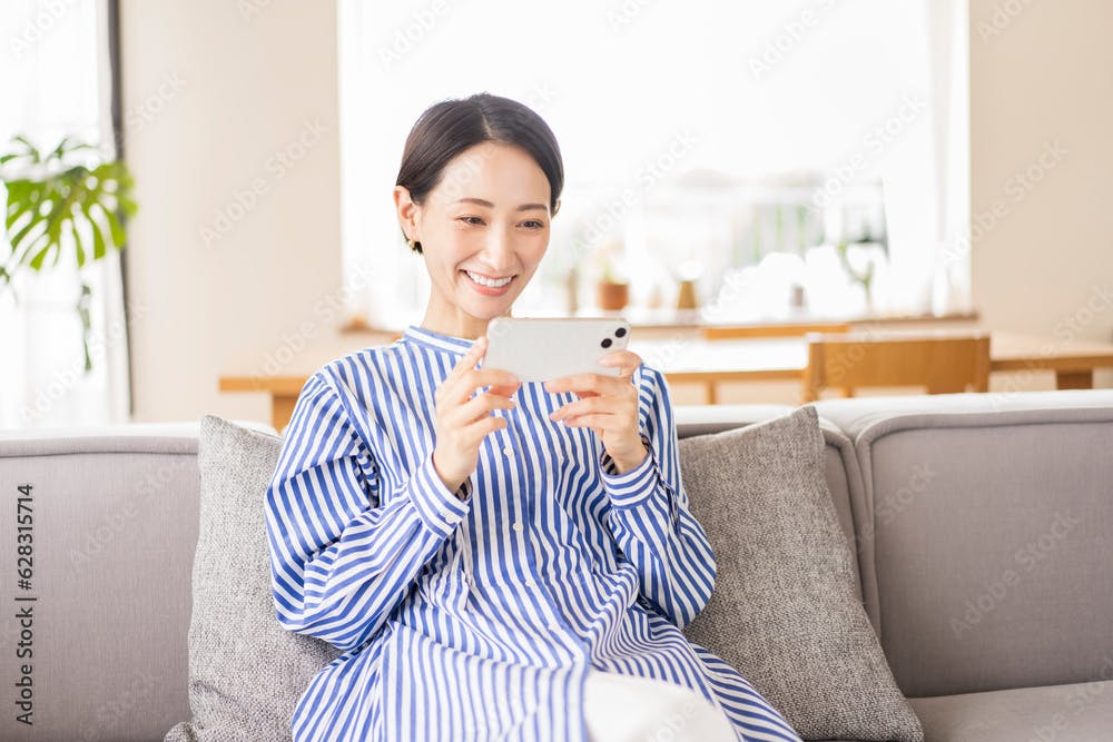 リビングでソファーに座りくつろぐ日本人ミドル女性/スマートフォンで動画を見る