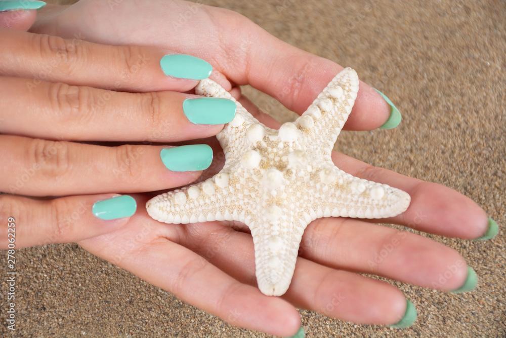 Young girl hands with a turquoise color nails polish holding starfish and sea sand in the background. Manicure and beauty concept. Close up, selective focus