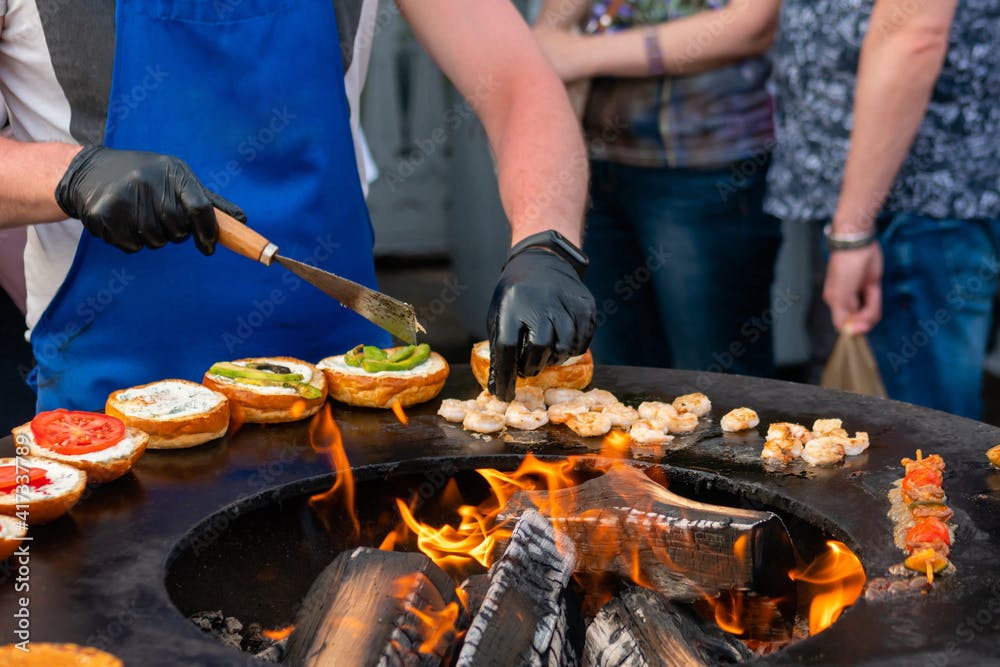 Chef preparing fish burgers with seafood, avocado, shrimp, prawn on brazier with hot flame at summer local food market - close up view. Outdoor cooking, gastronomy, cookery, street food concept
