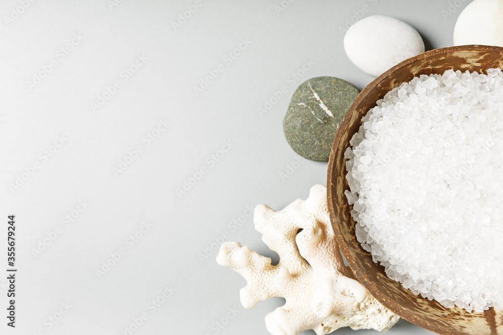  White sea bath salt in a coconut bowl, coral and stones on a gray background. Spa and relaxation concept. copy space